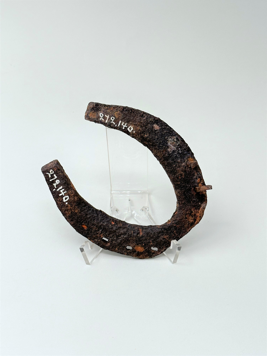 Hästsko i järn. Med fyra hål på vardera sida för söm. Två hål har kvarsittande spik. Kraftigt anfrätt av rost.