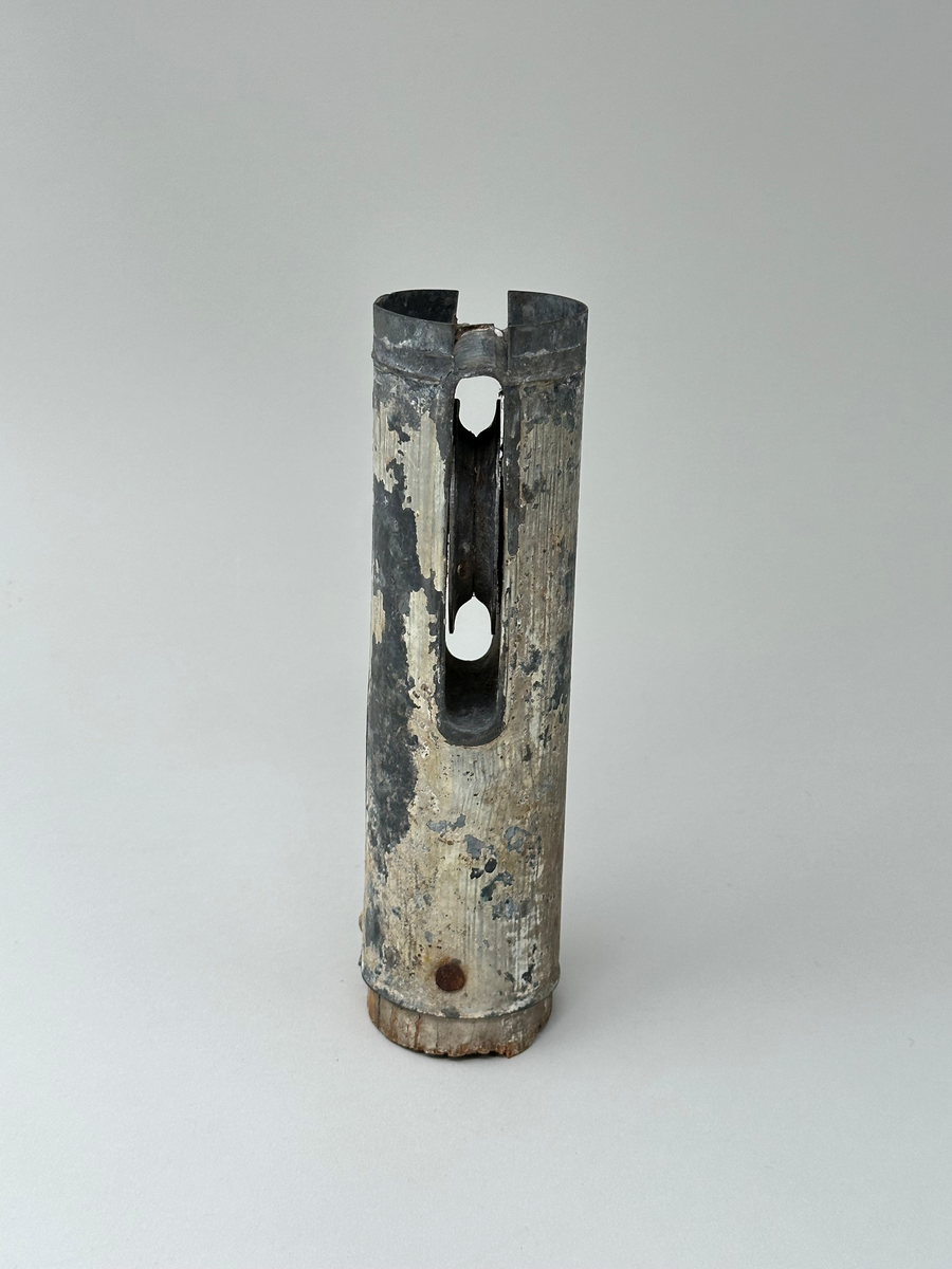 Del av flaggstång. Plåtcylinder sittande på övre delen av flaggstången med trissa för flagglina. Har varit vitmålad, spår av färg finns kvar. 