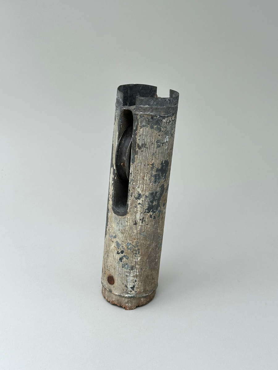 Del av flaggstång. Plåtcylinder sittande på övre delen av flaggstången med trissa för flagglina. Har varit vitmålad, spår av färg finns kvar. 