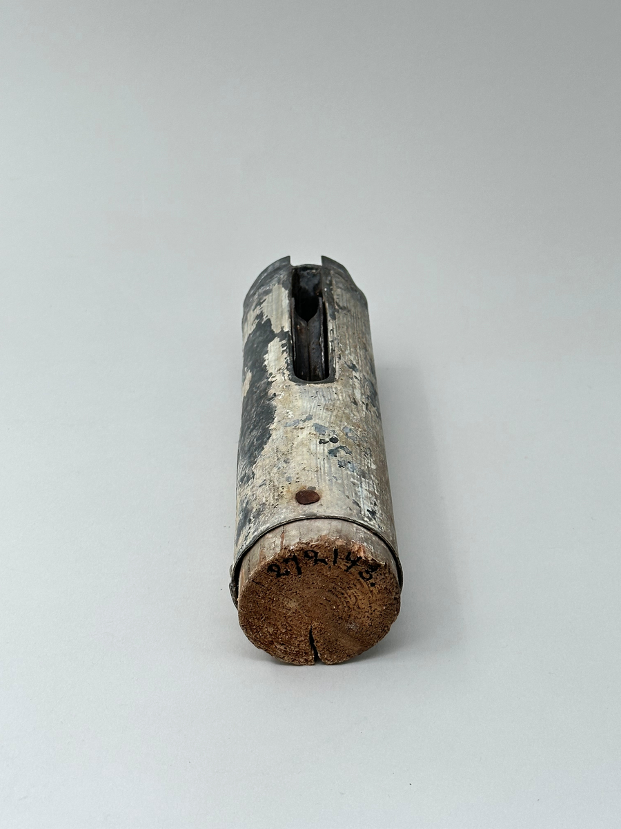 Del av flaggstång. Plåtcylinder sittande på övre delen av flaggstången med trissa för flagglina. Har varit vitmålad, spår av färg finns kvar. 