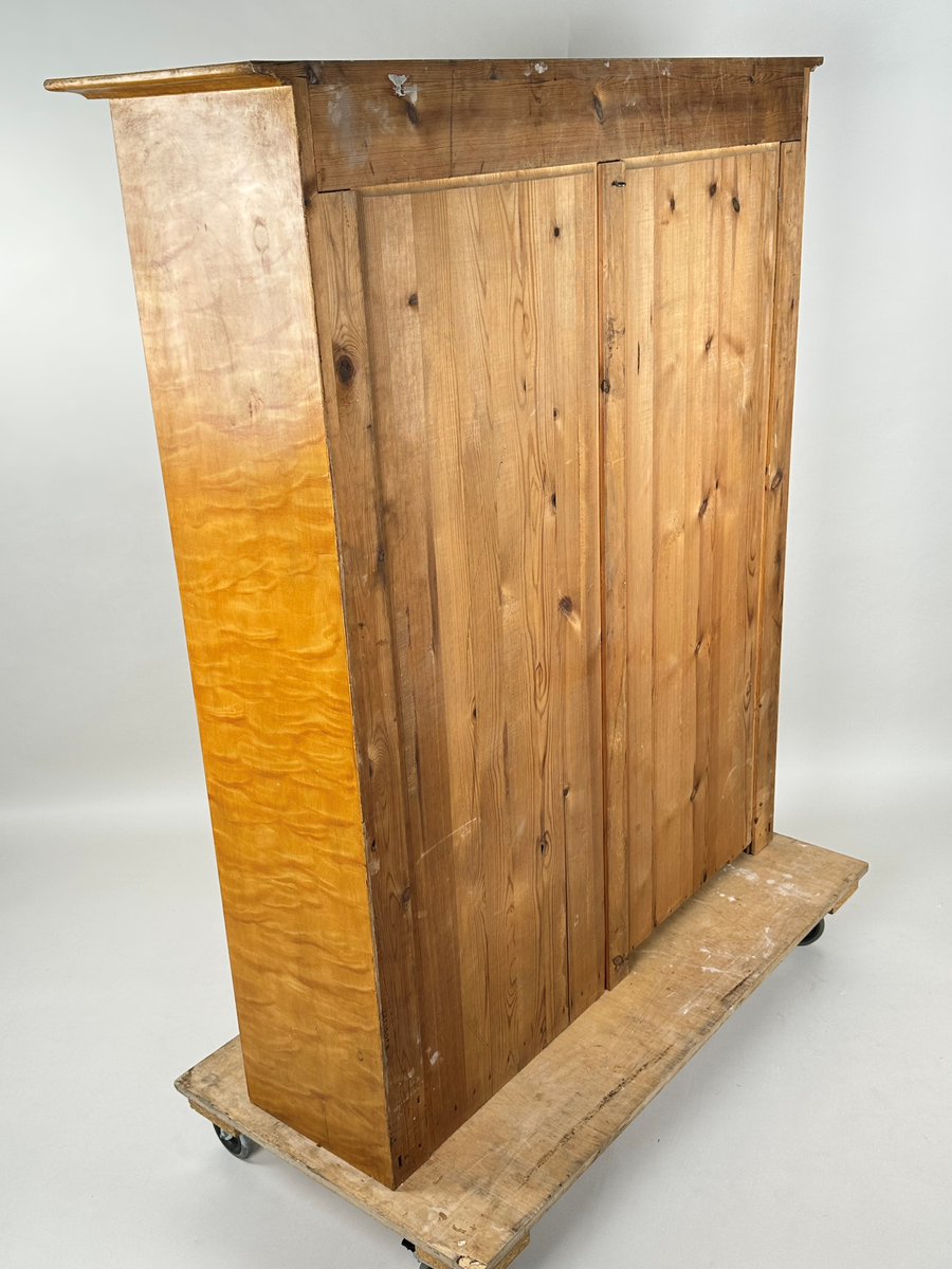 Materialskåp i trä, överskåp. Ådringsmålad i björkådring. Vitmålad på insidan. Två höga glasdörrar med två rutor vardera som skiljs åt vågrätt av en svängd list. De övre rutorna har en oval, något krusad överkant. Ena dörren med nyckelbricka i mässing. Räfflade fronter på skåpets sidostycken. Toppskiva profilerad undertill och överhängande.