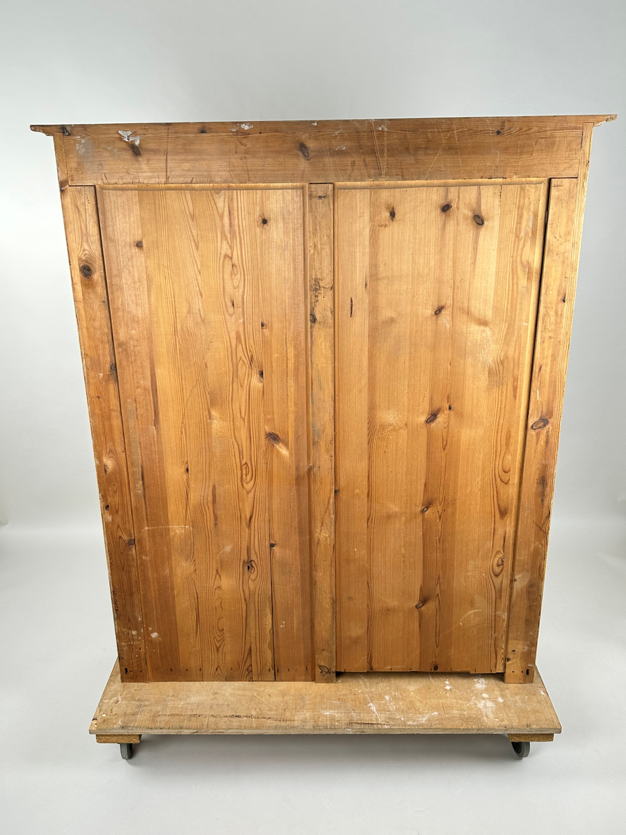 Materialskåp i trä, överskåp. Ådringsmålad i björkådring. Vitmålad på insidan. Två höga glasdörrar med två rutor vardera som skiljs åt vågrätt av en svängd list. De övre rutorna har en oval, något krusad överkant. Ena dörren med nyckelbricka i mässing. Räfflade fronter på skåpets sidostycken. Toppskiva profilerad undertill och överhängande.