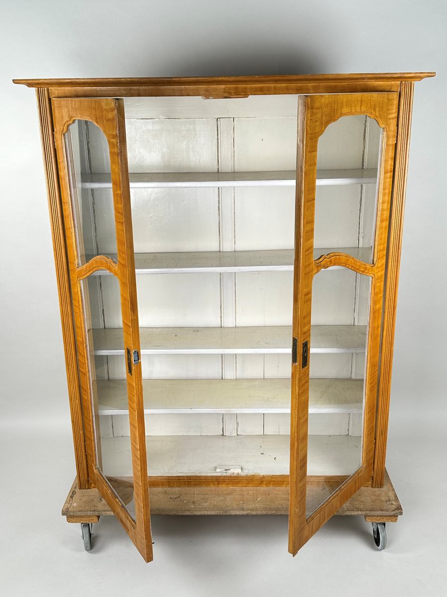 Materialskåp i trä, överskåp. Ådringsmålad i björkådring. Vitmålad på insidan. Två höga glasdörrar med två rutor vardera som skiljs åt vågrätt av en svängd list. De övre rutorna har en oval, något krusad överkant. Ena dörren med nyckelbricka i mässing. Räfflade fronter på skåpets sidostycken. Toppskiva profilerad undertill och överhängande.