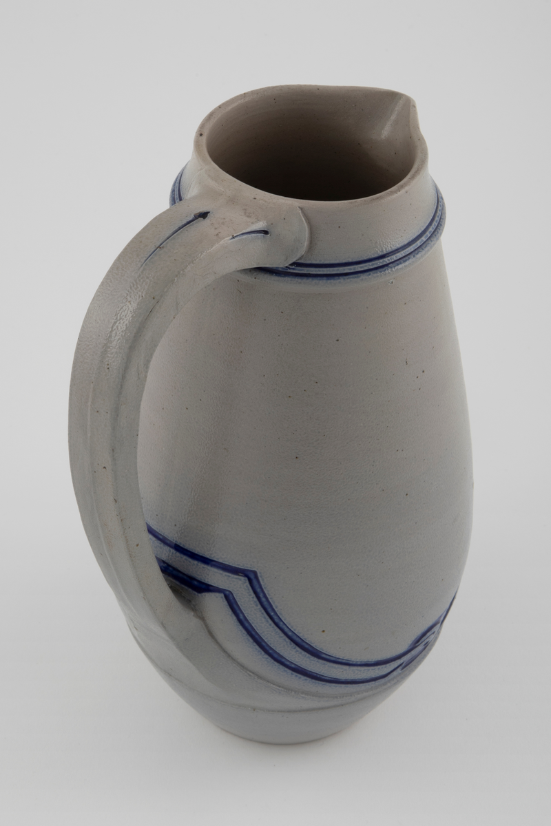 Kanne med grå bunn og koboltblå dekor. Kannen har stor buk. Dekoren er innskjært i leiren og fylt med koboltglasur. Dette gir effekten av dypt relieff. Jolly-dreid korpus. Håndtak er lagd separat. Kannen er glasert med saltglasur og dekoren med kobolt.