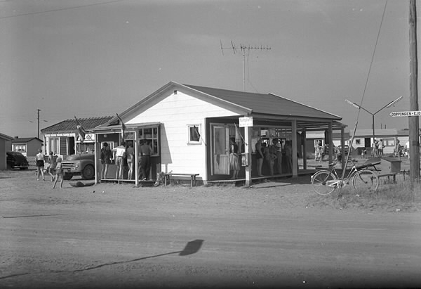 Pilängens livsmedel i Olofsbo där människor köar vid kioskluckan. Till höger bakom kiosken spelas det minigolf. Runtom ses stugor, parkerade fordon och en cykel. På en stolpe till höger sitter en skylt med texten: "Doppingen Ejdern".