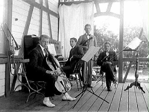 Orkester år 1919 på verandan till paviljongen på Gamla Påskberget. De spelar cello, piano och fioler. På väggen hänger även en trombon och till höger står ett notställ.