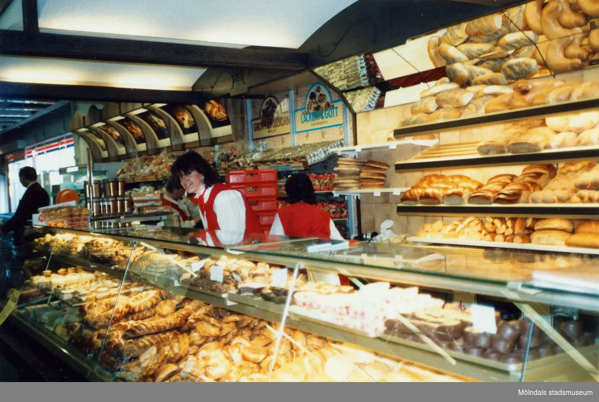 Två kvinnliga expediter klädda i röda rockar står innanför bröddisken, Brödavdelningen på K-marknad, Mölndals C efter 1985/86. Hyllorna är fyllda av allehanda bakverk.