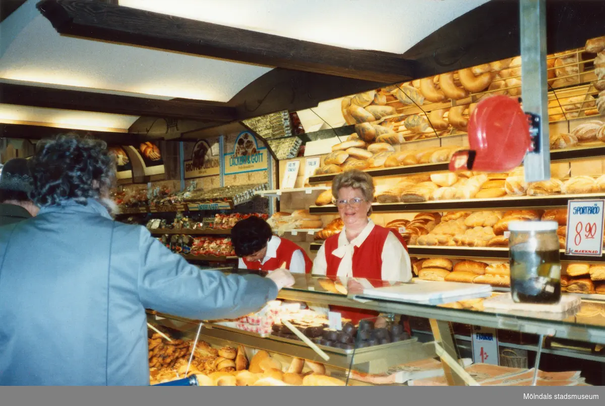 Två kvinnliga expediter klädda i röda rockar står innanför bröddisken, Brödavdelningen på K-marknad, Mölndals C efter 1985/86. Hyllorna är fyllda av allehanda bakverk och en kund håller fram sin hand. På en stolpe sitter en röd kölapps-apparat.
