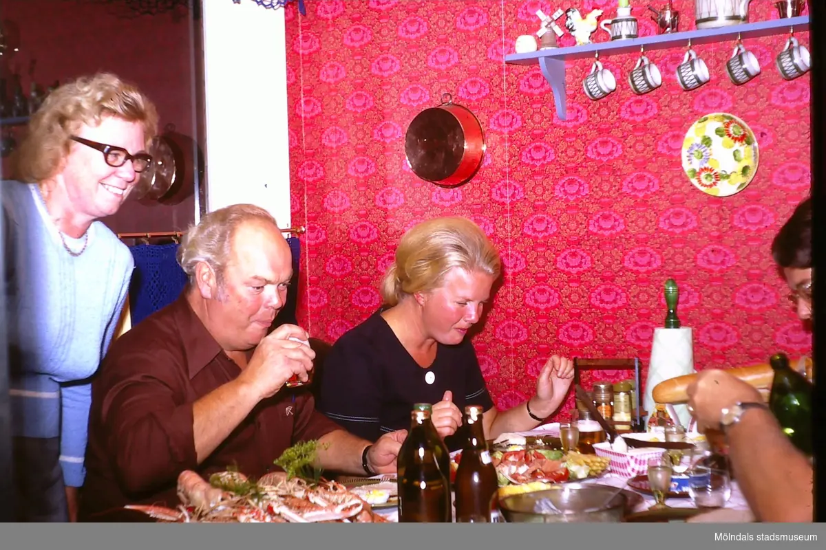 Kräftskiva i familjen Thörnqvists kök, Lindome okänt årtal. Berit Thörnqvist står längst till vänster. Vid köksbordet sitter tre vuxna personer och äter, Bordet står utmed en vägg, som har en starkt färgad tapet med cerice och orangefärgade blommor. Tidstypiska föremål sätter uppsatta på väggen.