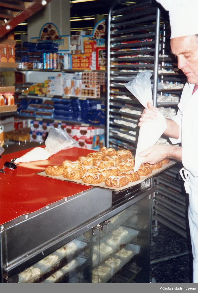 En manlig bagare klädd i vitt spritsar nybakade wienerbröd med sockerkristyr, K-marknad efter 1985/86. Till höger om honom står en ställning som innehåller flera plåtar med bakverk.