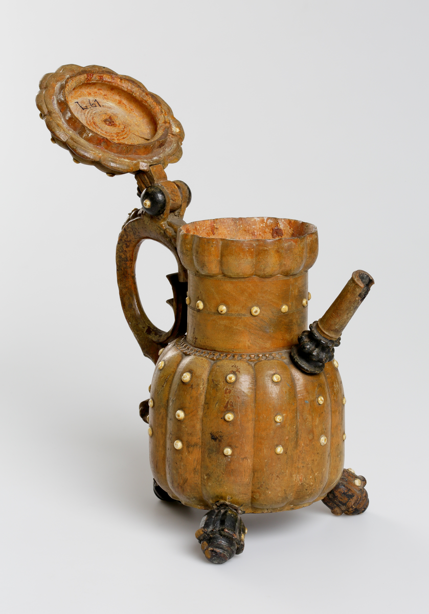 Svarvad och snidad kanna av masur med inläggningar av benknappar. Buken har vertikala åsar, indraget cylindriskt halsparti med utåtböjd bräddkant. Pipens nedre del är profilerad och den övre i det närmaste cylindrisk, vilken är fäst i kannan vid övergången till halsen. Locket är ledat vid handtaget med ett rosettformigt svartmålat mittparti. Handtaget är s-formigt med en skuren pärlstav på utsidan. Nedtill på buken finns tre stycken svartmålade och blomkalkformiga fötter. Runt hela kannan på buk, hals, lock, handtag och fötter sitter små runda benknappar. Mittknappen på lockets mitt är något större än övriga. 
Inskrivet i huvudkatalogen 1794.