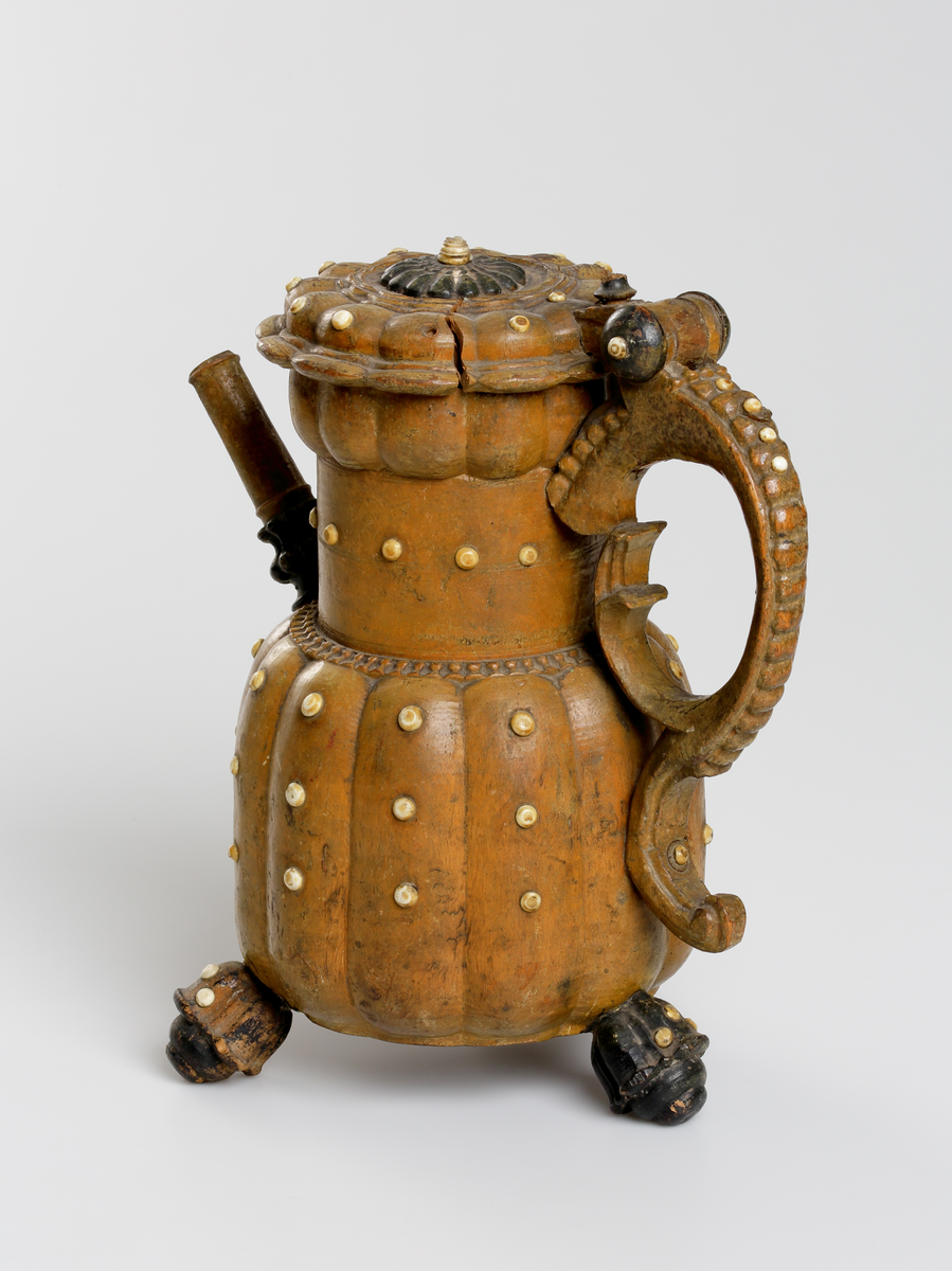 Svarvad och snidad kanna av masur med inläggningar av benknappar. Buken har vertikala åsar, indraget cylindriskt halsparti med utåtböjd bräddkant. Pipens nedre del är profilerad och den övre i det närmaste cylindrisk, vilken är fäst i kannan vid övergången till halsen. Locket är ledat vid handtaget med ett rosettformigt svartmålat mittparti. Handtaget är s-formigt med en skuren pärlstav på utsidan. Nedtill på buken finns tre stycken svartmålade och blomkalkformiga fötter. Runt hela kannan på buk, hals, lock, handtag och fötter sitter små runda benknappar. Mittknappen på lockets mitt är något större än övriga. 
Inskrivet i huvudkatalogen 1794.