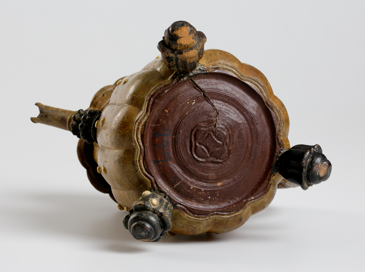 Svarvad och snidad kanna av masur med inläggningar av benknappar. Buken har vertikala åsar, indraget cylindriskt halsparti med utåtböjd bräddkant. Pipens nedre del är profilerad och den övre i det närmaste cylindrisk, vilken är fäst i kannan vid övergången till halsen. Locket är ledat vid handtaget med ett rosettformigt svartmålat mittparti. Handtaget är s-formigt med en skuren pärlstav på utsidan. Nedtill på buken finns tre stycken svartmålade och blomkalkformiga fötter. Runt hela kannan på buk, hals, lock, handtag och fötter sitter små runda benknappar. Mittknappen på lockets mitt är något större än övriga. 
Inskrivet i huvudkatalogen 1794.