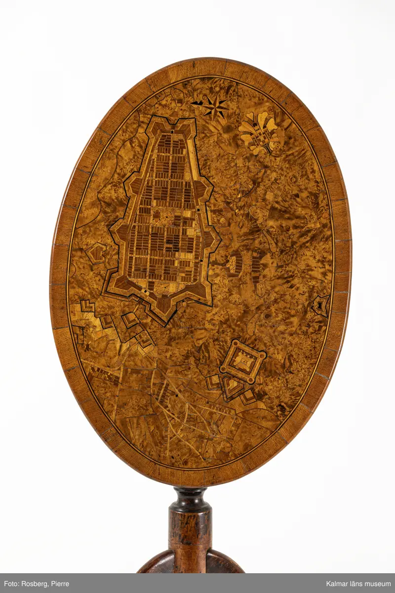 KLM 10594. Bord, fällbord. Av trä. Oval skiva av alrot med rika inläggningar, återgivande stadsplan över Kalmar stad med  Kvarnholmen och Kalmar slott, Gamla stadens gatunät, Grimskär skans, inläggning med norrpil samt Stadsporten som ett dekorativt element med blad. Pelare, svarvad och profilerad med tredelad fot. Fällbar bordsskiva med mekanism som fungerar. Datering, gustaviansk, 1700-talet slut.