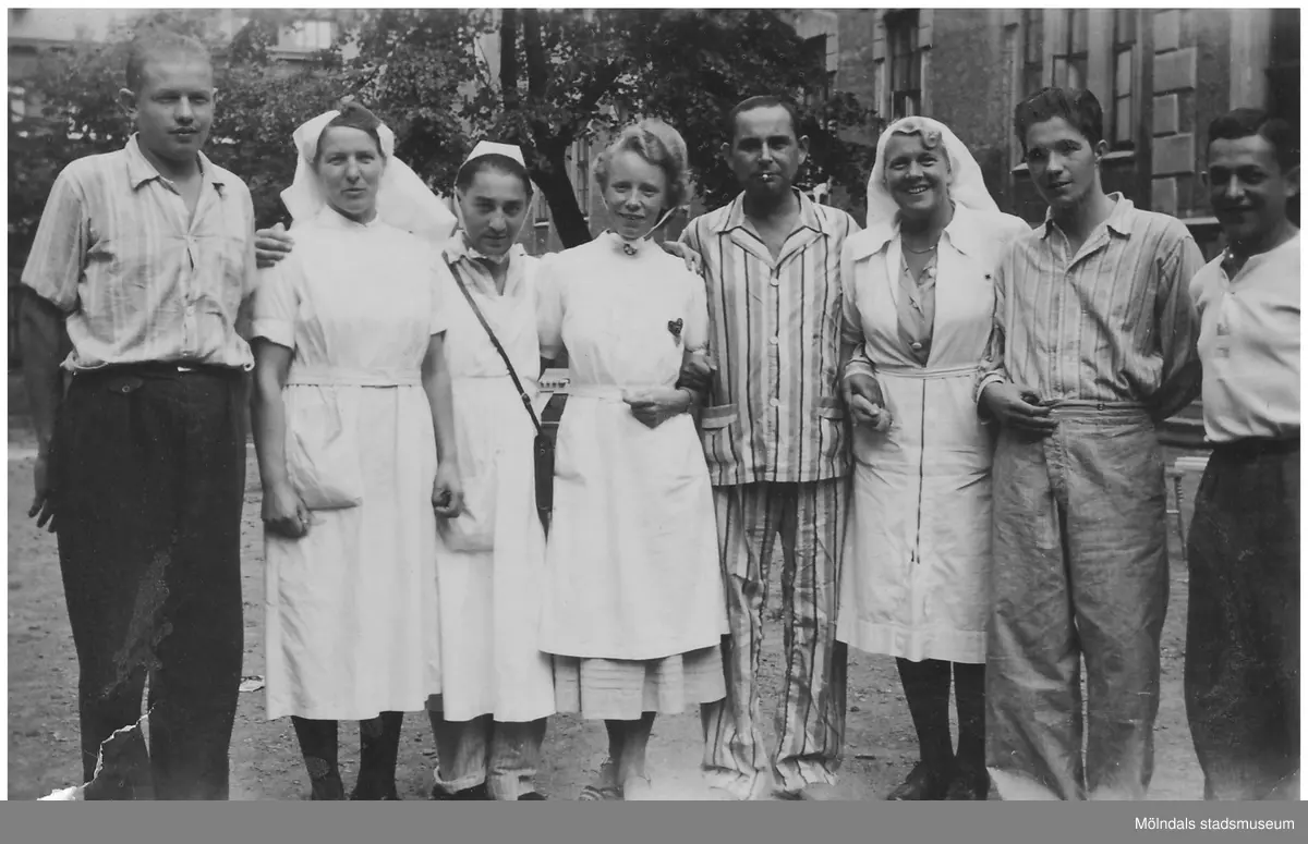 "Karantänsförläggning" på Kvarnbyskolan för överlevare från koncentrationsläger i Tyskland och Polen 1945. Skolan fungerade under denna sommar som beredsskapssjukhus.
Åtta personer står bredvid varandra utanför Kvarnbyskolan. Det är fem män (flyktingar) och tre sjuksköterskor. Femte personen från vänster är en man klädd i pyjamas som håller två sköterskor i armarna. Mannen mellan de två sköterskorna skojar genom att vara utklädd till sjuksköterska. Längst till höger ses Abraham Frischer (1923 - 2009).
Relaterade motiv: 2024_1281 - 1299.