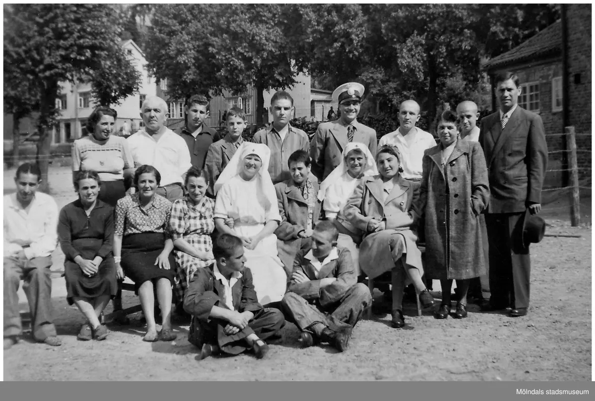 "Karantänsförläggning" på Kvarnbyskolan för överlevare från koncentrationsläger i Tyskland och Polen 1945. Skolan fungerade under denna sommar som beredsskapssjukhus.
Tjugo personer sitter/står utanför Kvarnbyskolan. Det är en blandning av män och kvinnor varav två är sjuksköterskor. En man är klädd i uniform. Några personer ler och några ser allvarliga ut. Byggnader i bostadsområdet Trädgården ses i bakgrunden.