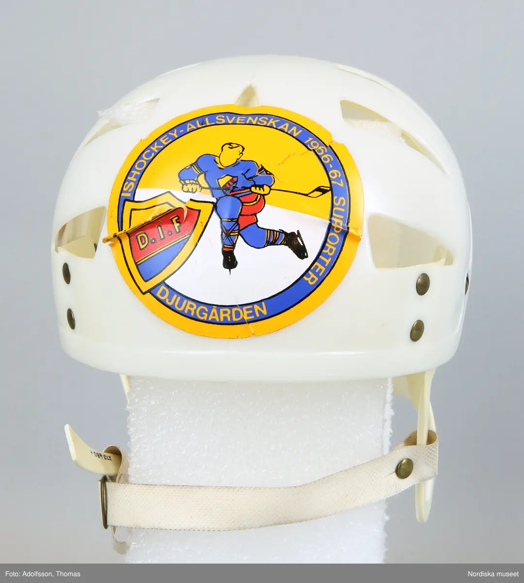 Inventering Sesam 1996-1999:
L  21   H  19 (cm)
Ishockeyhjälm, av gjuten vit hårdplast, genombruten, hopsatt med nitar av gulmetall, ¿ronbyglar med reglerbart hakband av plastad, kraftig väv med tryckknapp, ingjutet baktill: "ABC-FABRIKERNA / KUNGÄLV SWEDEN". 2 st påklistrade etiketter; r¿d etikett med vit text: "UFFE" och rund etikett med bild av ishockeyspelare samt sk¿ld med text: "D.I.F." och "ISHOCKEYALLSVENSKAN 1966-67 SUPPORTER  DJURGÅRDEN".
En bygel trasig.
Anna Womack mars 1998