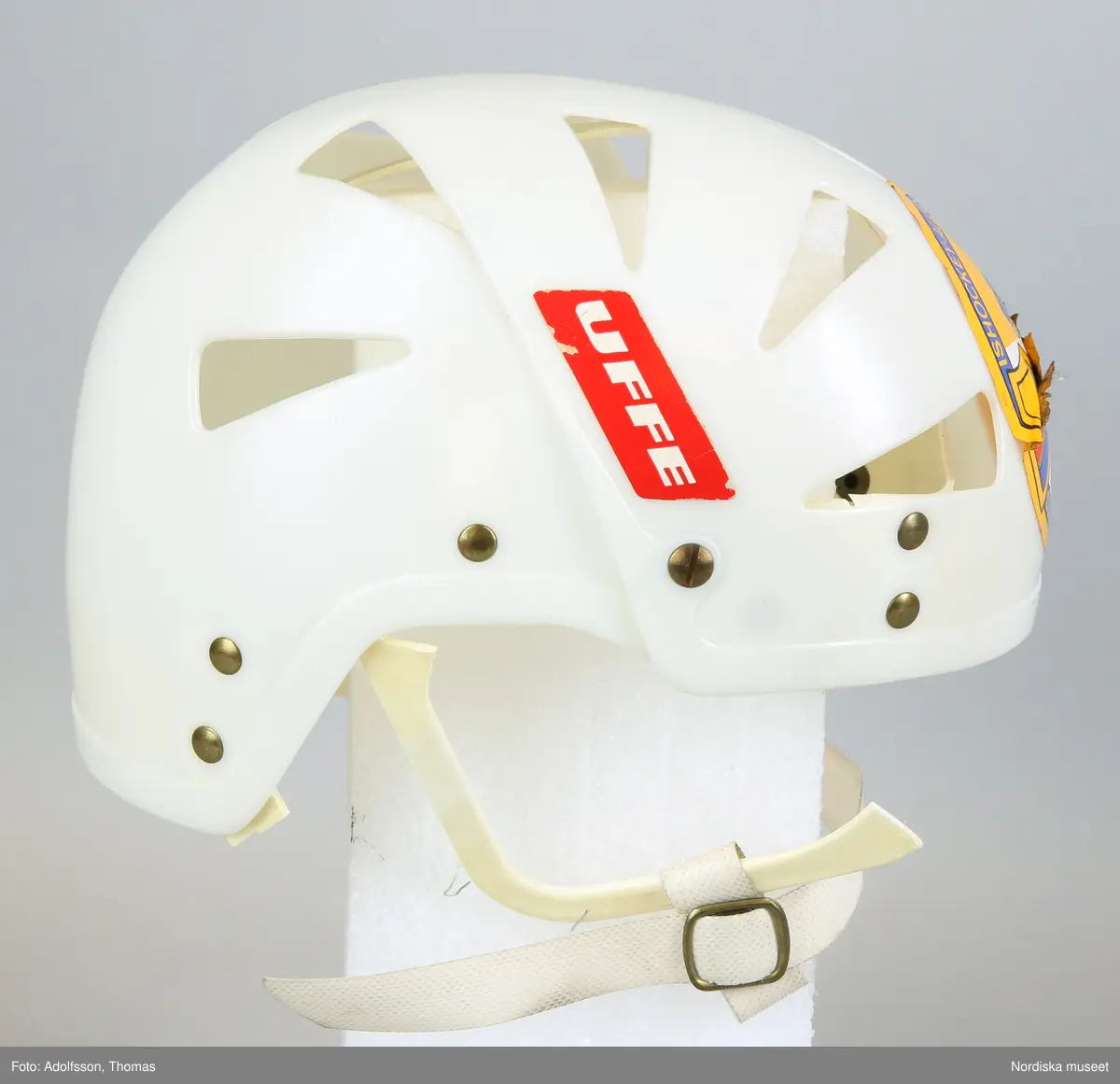 Inventering Sesam 1996-1999:
L  21   H  19 (cm)
Ishockeyhjälm, av gjuten vit hårdplast, genombruten, hopsatt med nitar av gulmetall, ¿ronbyglar med reglerbart hakband av plastad, kraftig väv med tryckknapp, ingjutet baktill: "ABC-FABRIKERNA / KUNGÄLV SWEDEN". 2 st påklistrade etiketter; r¿d etikett med vit text: "UFFE" och rund etikett med bild av ishockeyspelare samt sk¿ld med text: "D.I.F." och "ISHOCKEYALLSVENSKAN 1966-67 SUPPORTER  DJURGÅRDEN".
En bygel trasig.
Anna Womack mars 1998