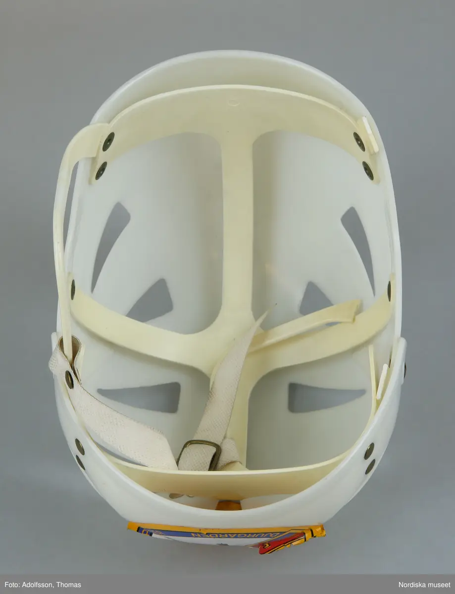 Inventering Sesam 1996-1999:
L  21   H  19 (cm)
Ishockeyhjälm, av gjuten vit hårdplast, genombruten, hopsatt med nitar av gulmetall, ¿ronbyglar med reglerbart hakband av plastad, kraftig väv med tryckknapp, ingjutet baktill: "ABC-FABRIKERNA / KUNGÄLV SWEDEN". 2 st påklistrade etiketter; r¿d etikett med vit text: "UFFE" och rund etikett med bild av ishockeyspelare samt sk¿ld med text: "D.I.F." och "ISHOCKEYALLSVENSKAN 1966-67 SUPPORTER  DJURGÅRDEN".
En bygel trasig.
Anna Womack mars 1998