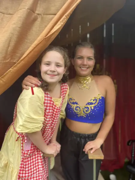 To jenter ikledd kostymer smiler til kamera. En er utkledd som sirkusartist, en er utkledd i gul kjole med rød-rutete forkle.