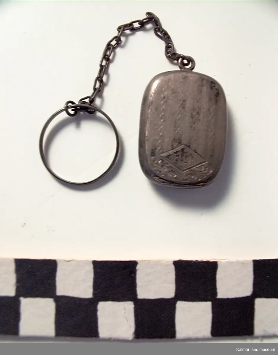 KLM 40369:4 Medaljong, metall, fäst vid kort kedja med en liten ring i andra änden. På utsidan initialerna AB, inuti en stämpel. Kan eventuellt vara en liten ask.