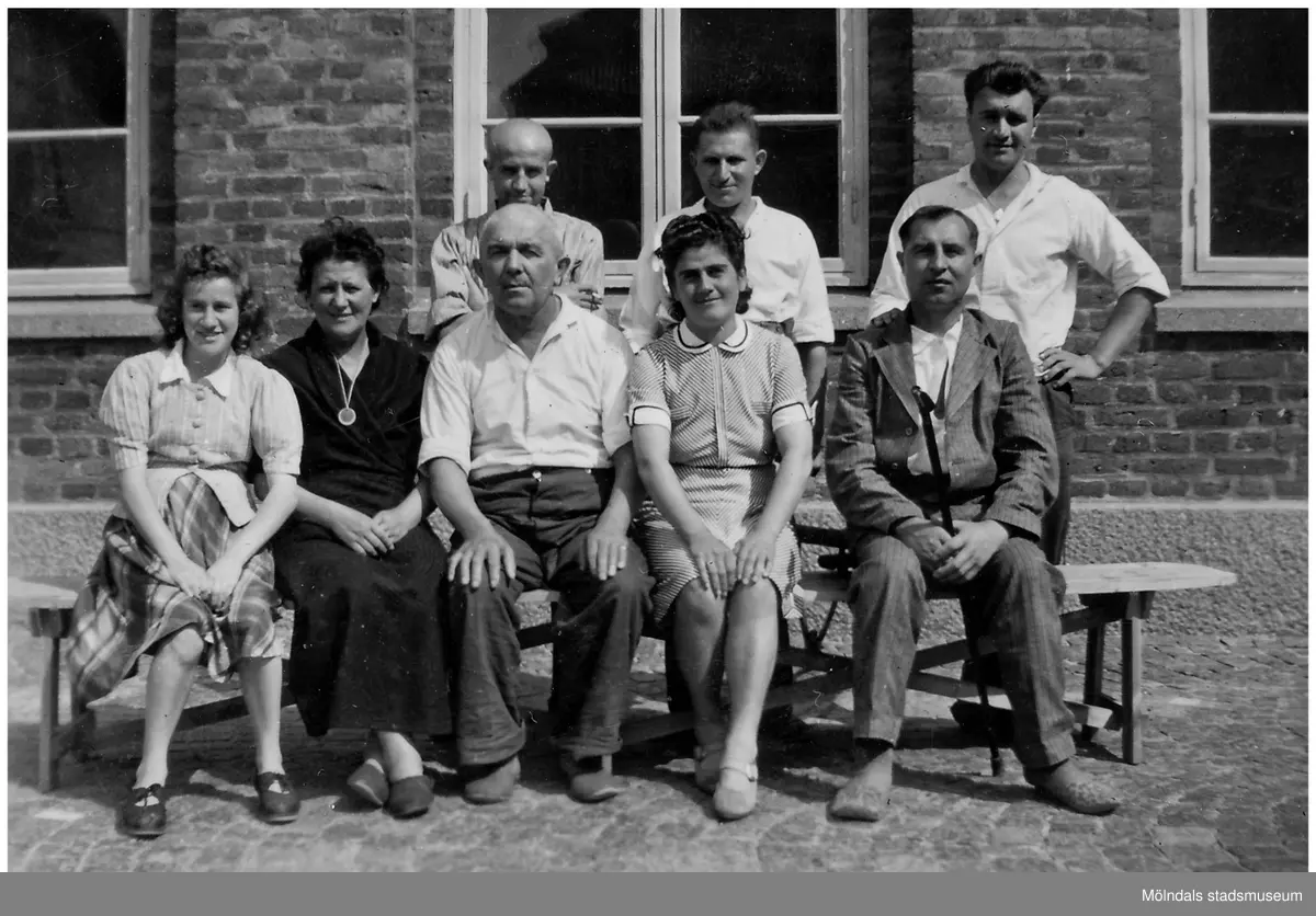"Karantänsförläggning" på Kvarnbyskolan för överlevare från koncentrationsläger i Tyskland och Polen 1945. Skolan fungerade under denna sommar som beredsskapssjukhus.
Åtta personer befinner sig utanför Kvarnbyskolan. Tre kvinnor kvinnor och två män sitter på en träbänk. Tre män står bakom dem.
