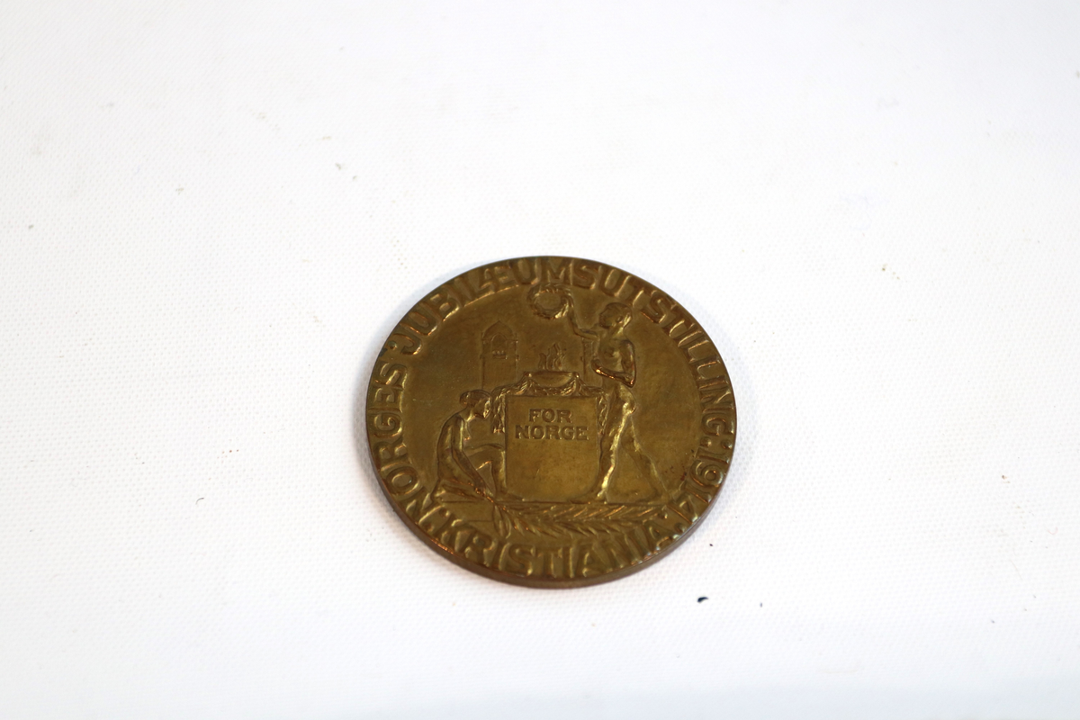Medalje i metall. Rund form. Det norske riksvåpen omkretset av en løvtregrein på hver side, på den ene siden, og en kvinne og en mann i profil på andre siden. Mannen holder en krans over en ild, kvinnen sitter på venstre side og holder en stor gren som ligger på bakken.