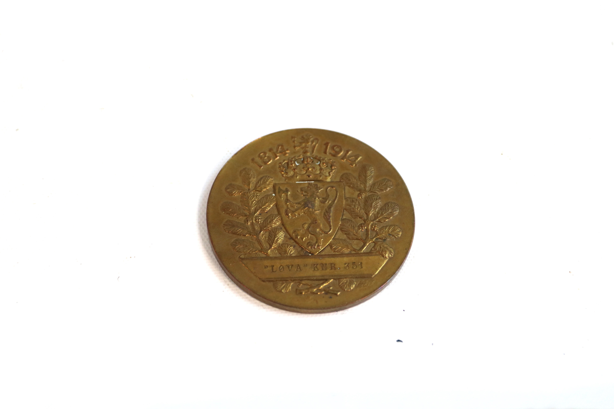 Medalje i metall. Rund form. Det norske riksvåpen omkretset av en løvtregrein på hver side, på den ene siden, og en kvinne og en mann i profil på andre siden. Mannen holder en krans over en ild, kvinnen sitter på venstre side og holder en stor gren som ligger på bakken.