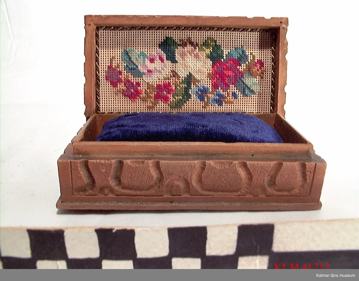 KLM 41213 Ask, smyckeskrin, trä, textil. Liten rektangulär ask med fällock, av snidat trä. På ovansidan snidade blommor. Inuti är locket klätt med tyg med broderade blommor i rött, blått och vitt. Botten är klädd med en blå tygbit. I asken ligger en blå sammetskudde, troligen hemmagjord. Använd som smyckeskrin.