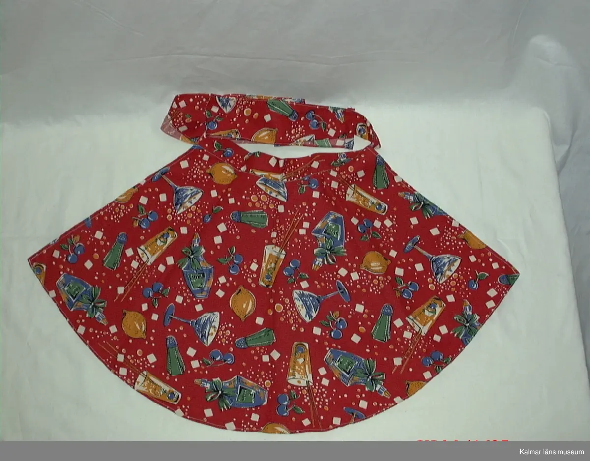KLM 41627 Förkläde, midjeförkläde, textil. Kort midjeförkläde av tyg med tryckt mönster, drinkglas, flaskor, frukter m m i blått, gult, grönt och vitt på röd botten, rundad nederkant, linning och knytband i ett av samma tyg.