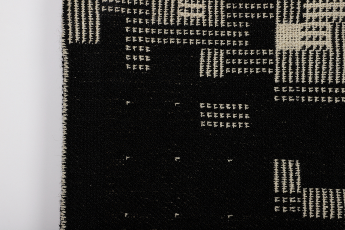 Rektangulær billedvev i svart og naturhvit ull. Komposisjonen er bygget opp som et digitalt bilde bestående av små kvadrater (piksel) med ulik mønster. Sett fra avstand danner disse et motiv av boligblokker.