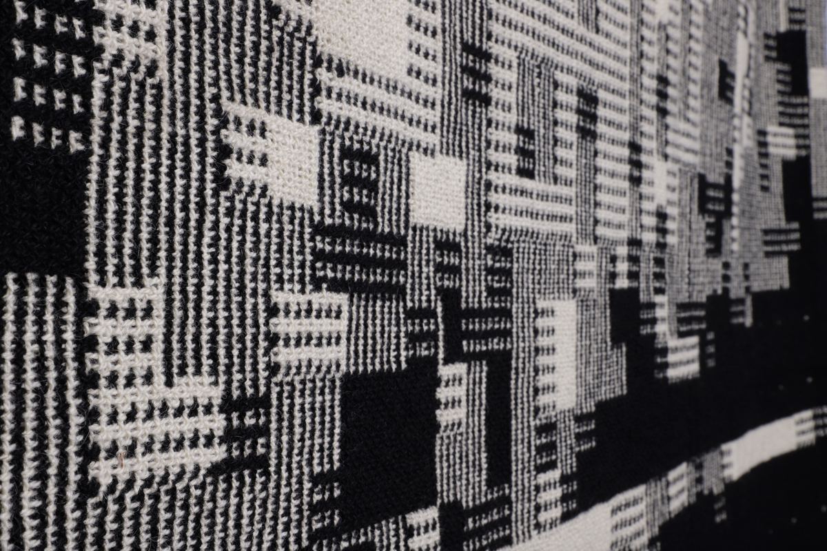Rektangulær billedvev i svart og naturhvit ull. Komposisjonen er bygget opp som et digitalt bilde bestående av små kvadrater (piksel) med ulik mønster. Sett fra avstand danner disse et motiv av boligblokker.