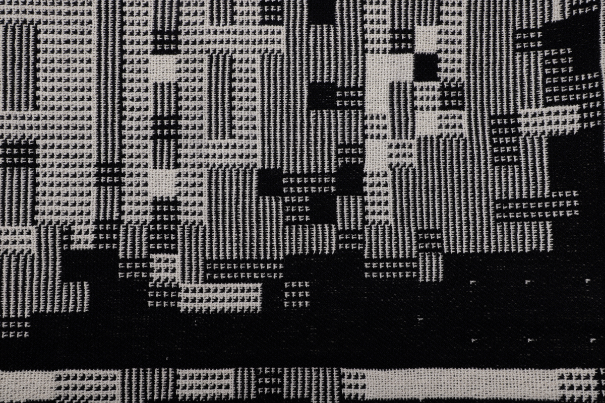 Rektangulær billedvev i svart og naturhvit ull. Komposisjonen er bygget opp som et digitalt bilde bestående av små kvadrater (piksel) med ulik mønster. Sett fra avstand danner disse et motiv av boligblokker.