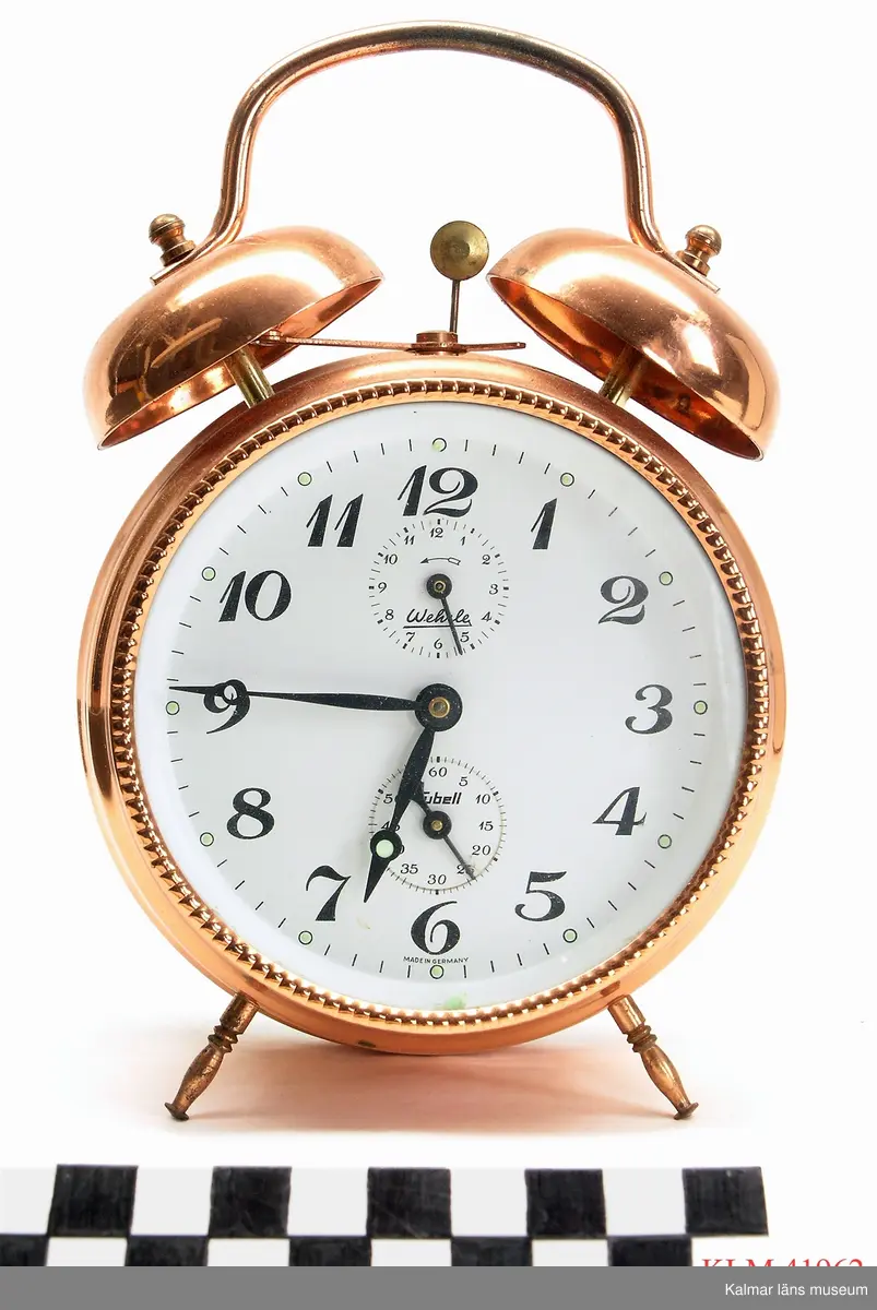 KLM 41962 Väckarur, metall. Mekaniskt. Runt ur av kopparfärgad metall, två små vinklade ben. Vit urtavla med siffrorna 1 till 12, tim- och minutvisare samt två mindre cirklar med visare för sekunder respektive väckningstid. Upptill väckningsfunktionen, en liten hammare som slår mot två klockor. Ett handtag är fäst i toppen på klockorna.