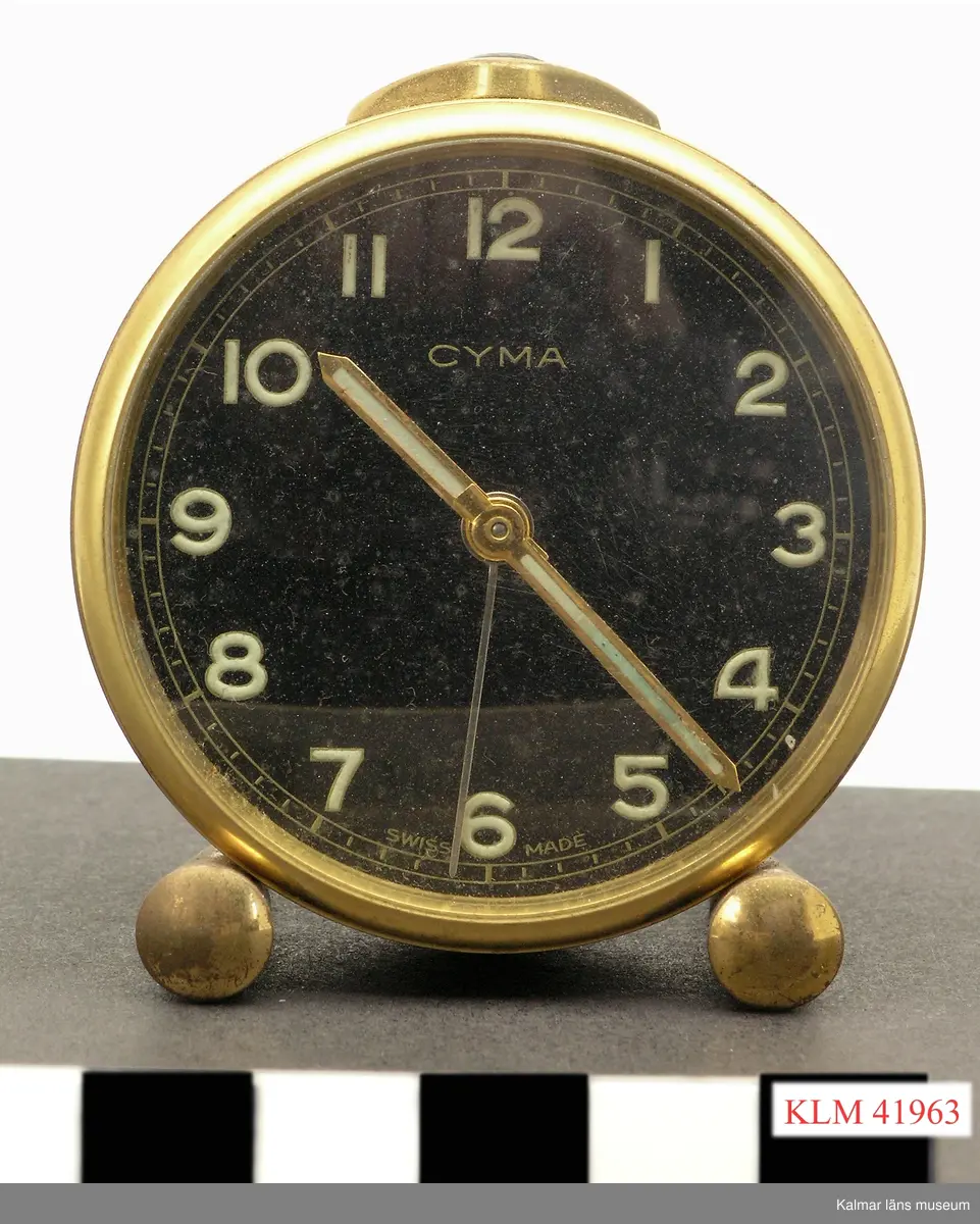 KLM 41963 Väckarur, metall. Mekaniskt. Runt ur av mässingfärgad metall, vilar på två fötter. Svart urtavla med siffrorna 1 till 12, tim- och minutvisare samt en tunnare visare för sekunder eller för väckningstid. Text "CYMA" samt "SWISS MADE". Upptill knapp för avstägning av väckningssignal.