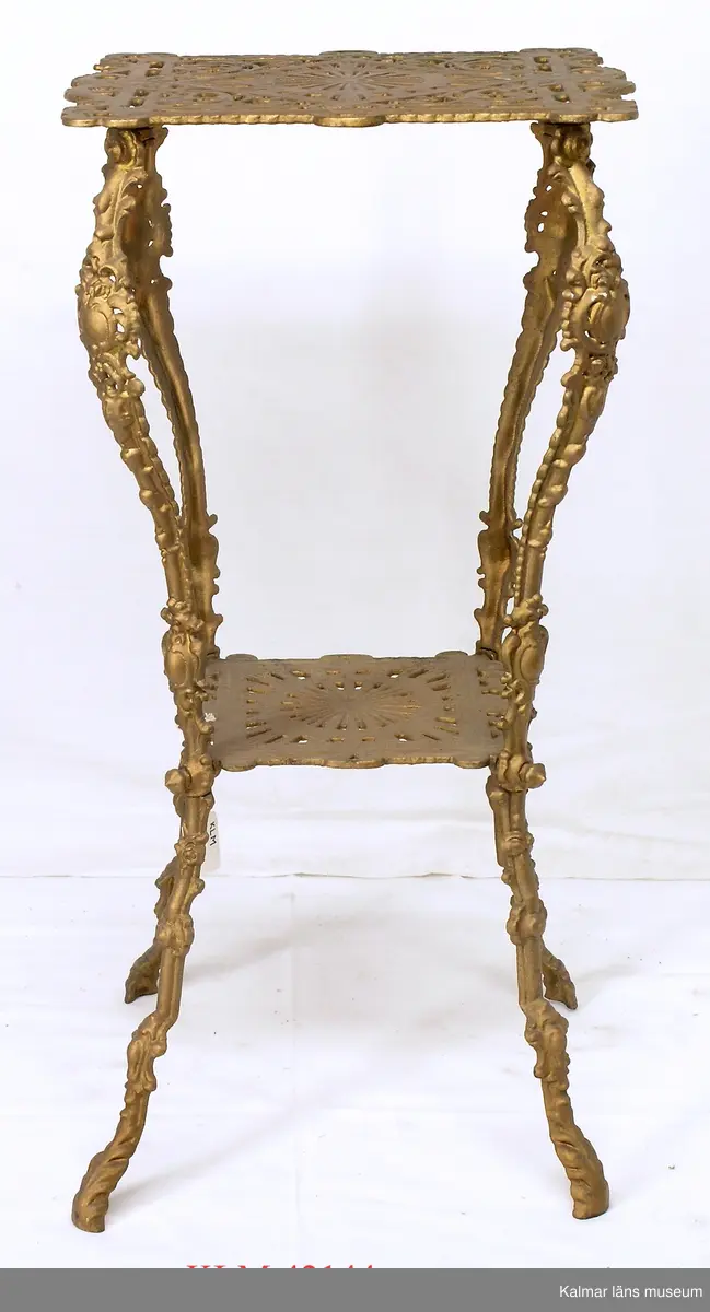 KLM 42144 Piedestal, gjutjärn. Kvadratisk skiva vilande på fyra svängda ben, där avståndet mellan benen är som minst sitter ytterligare en kvadratisk skiva. Hela piedestalen rikt ornamneterad och förgylld.