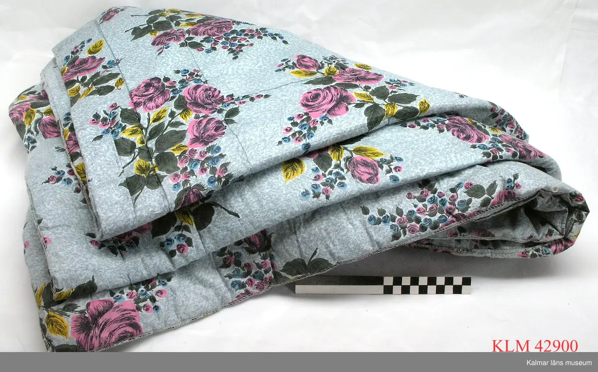 KLM 42900 Sängtäcke, textil. Rektangulärt täcke av tyg med tryckt mönster, rosenbuketter i rosa, blått, grönt och gult mot en småmönstrad ljusblå botten. Stoppat, stoppningen nedsydd med glesa linjära sömmar.