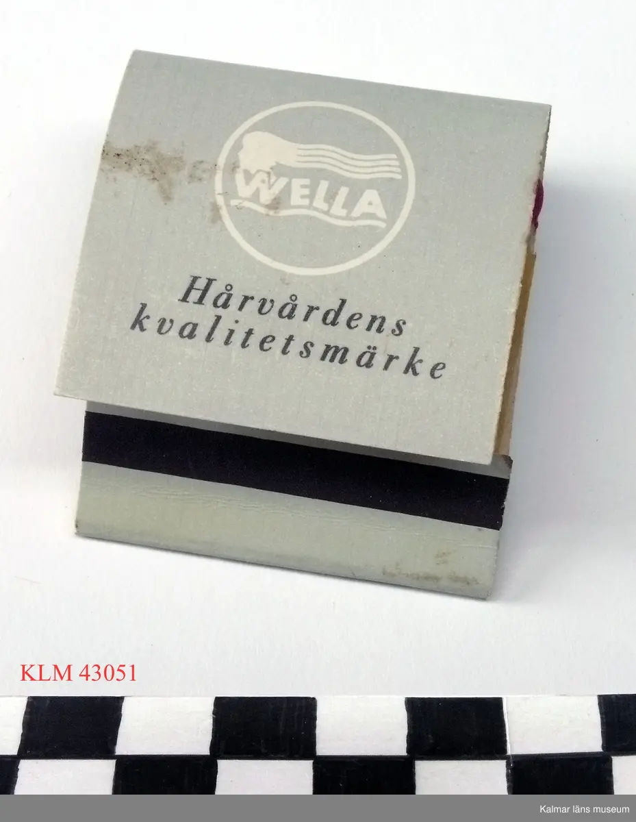 KLM 43051 Tändsticksask, tändsticksplån, papper. Rektangulärt, vikt av halvstyvt papper, grå utsida, tändplån, platta tändstickor av trä. Utsidan med reklamtext  för Wella Optawell kallpermanent, inuti tryckt text "M. Backmans Damfrisering Ö. Kyrkogatan 20 tel. 109 89".