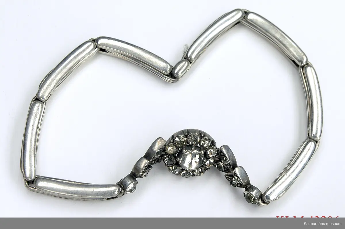 KLM 43206 Armband, silver, strass. Länkar av vit metall (silver?) förenade med töjbar tråd. Mellan två av länkarna är monterad en blomma av silver med infattade strasstenar. På blommans baksida stämpel "925" med en symbol under.