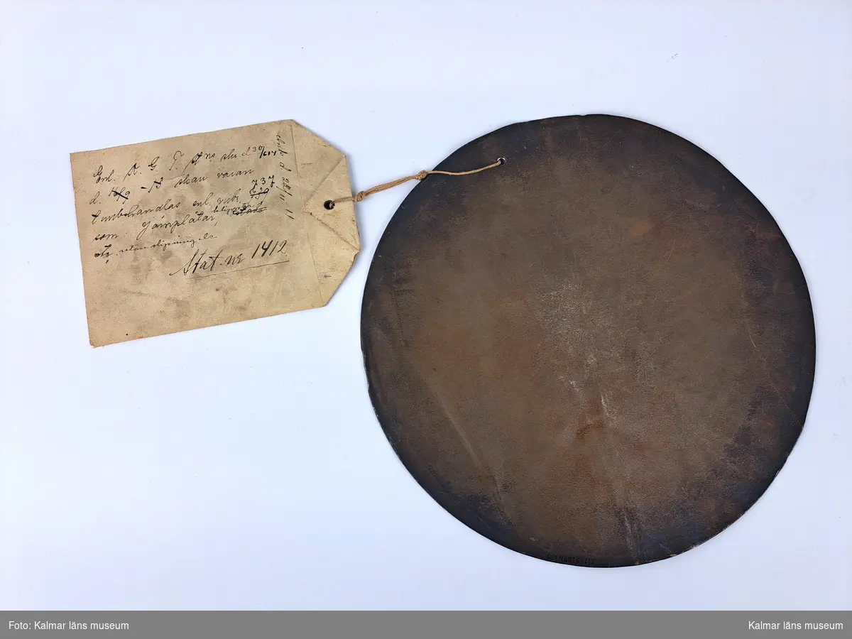 KLM 46816:1:1-2. Varuprov, järnplåt, 2 st. Två runda järnplåtar. :1. Med lapp i snöre, fäst genom hål i plåten. På lappen tryckt och handskriven text i svart på framsidan, handskriven text på baksidan samt en röd sigillstämpel. Text stämpel: Kongl Generaltullstyrelsen. På lappen handskrivet nummer: Stat. nr 1412. Påskrift plåt: ? 9157 1911. :2. En påklistad vit etikett samt påskrift plåt: Stat.nr.