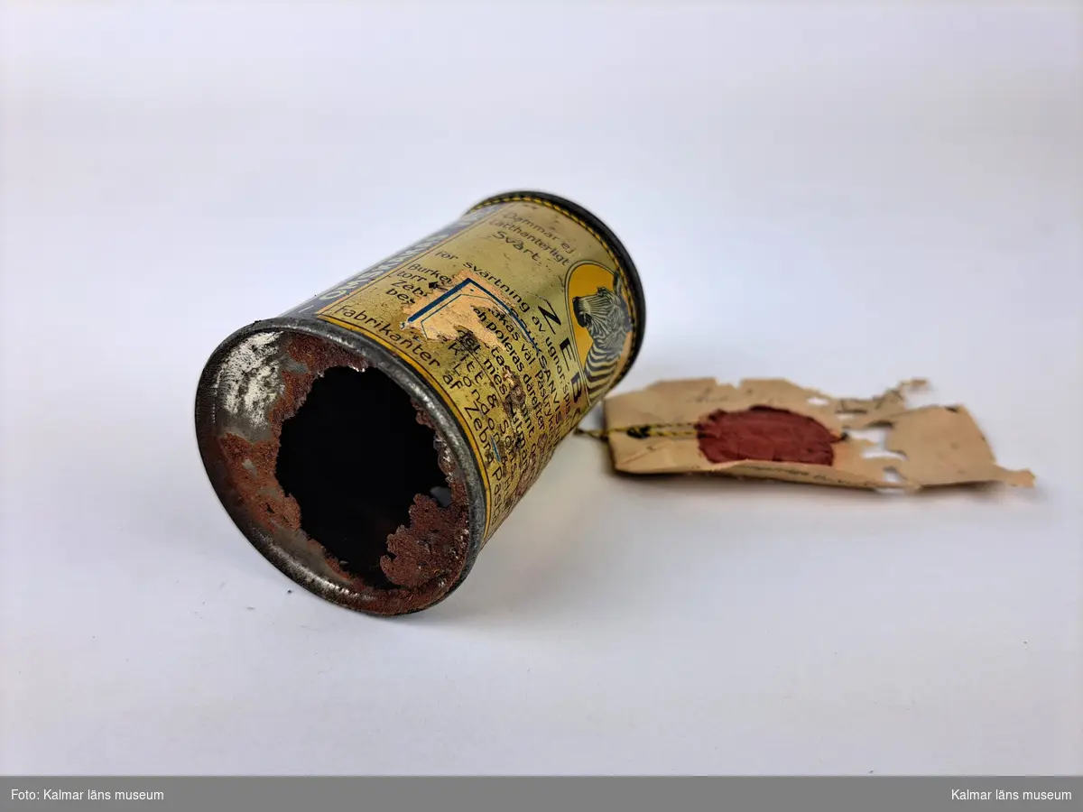 KLM 46816:16. Varuprov, förpackning. Rund metallburk med färgtryck och bild på en zebra. Trasig botten. Text: Zebo Flytande ugnssvärta, För svärtning av ugnar, spisar, järnstaket etc. Vit lapp/etikett, delvis trasig, hängande i textiltråd med tryckt och handskriven text (delvis otydlig): Stat. 603, Gust. T? Bratt & Co Göteborg (troligtvis). Handskriven text på baksidan samt en röd sigillstämpel. Text stämpel: Kongl Generaltullstyrelsen.