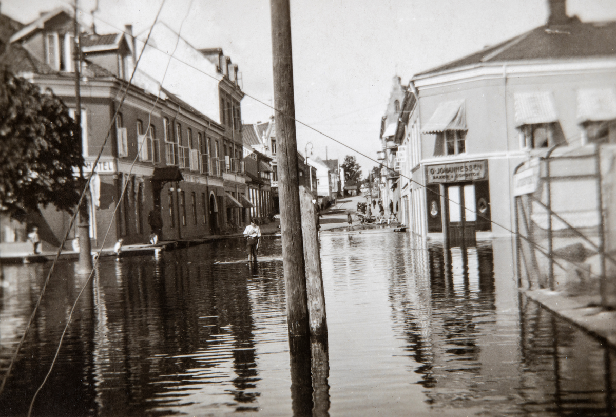 Stor flommen i Mjøsa juli 1927, Standgata står under vann. Bilde tatt fra jernbaneundergangen ved Viktoria hotell. Baker O. Johannessen sin foretning til høyre. Gutt som vasser i flomvannet.