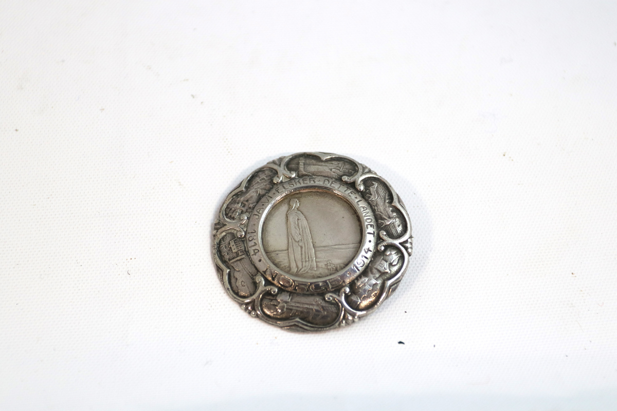 Medalje eller nål i sølv. Figur i profil i midten, med små, brede, nærmest hjerteformede felter, seks stykker. Alle med norske motiver. Sirkel i midten med figur på fremsiden har norske riksvåpen på baksiden og kan tas av. Ser ut til å være en jubileumsmynt.