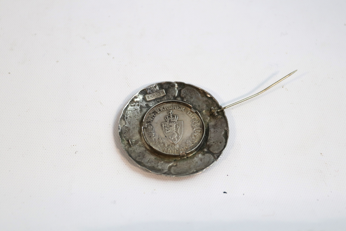 Medalje eller nål i sølv. Figur i profil i midten, med små, brede, nærmest hjerteformede felter, seks stykker. Alle med norske motiver. Sirkel i midten med figur på fremsiden har norske riksvåpen på baksiden og kan tas av. Ser ut til å være en jubileumsmynt.