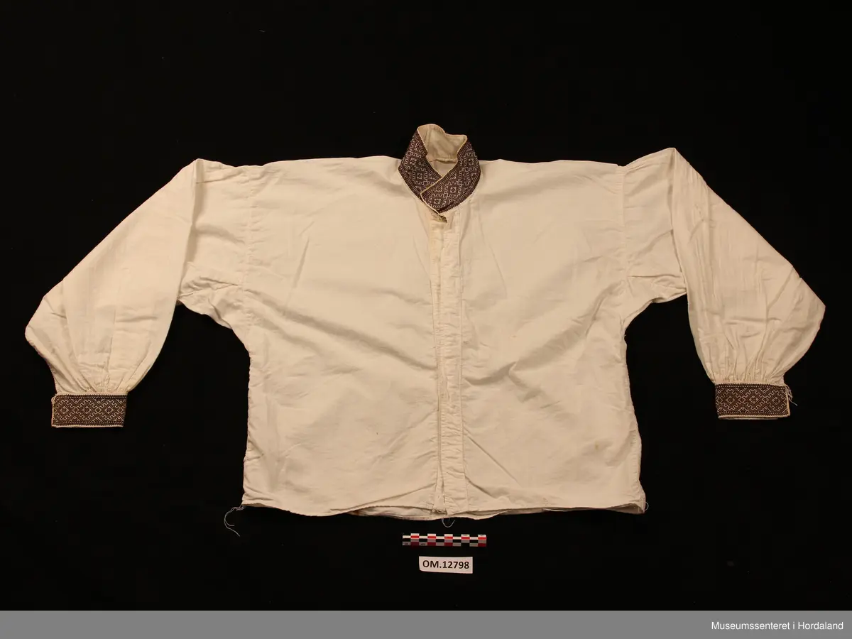 Bunadskjorte i bomullslerret med halskrage og handlinningar i smøyg (broderi). Brodert på svært tynt bomullstoff. Skjorta har isydde ermer med litt rynker på ermetoppen. Framme på ermene er det og rynker. Knapp og hempe på kvar handlinning. Stort knapphol på kvar side i halslinningen. Skjorta ber preg av at broderiet er eldre enn sjølve skjorta.