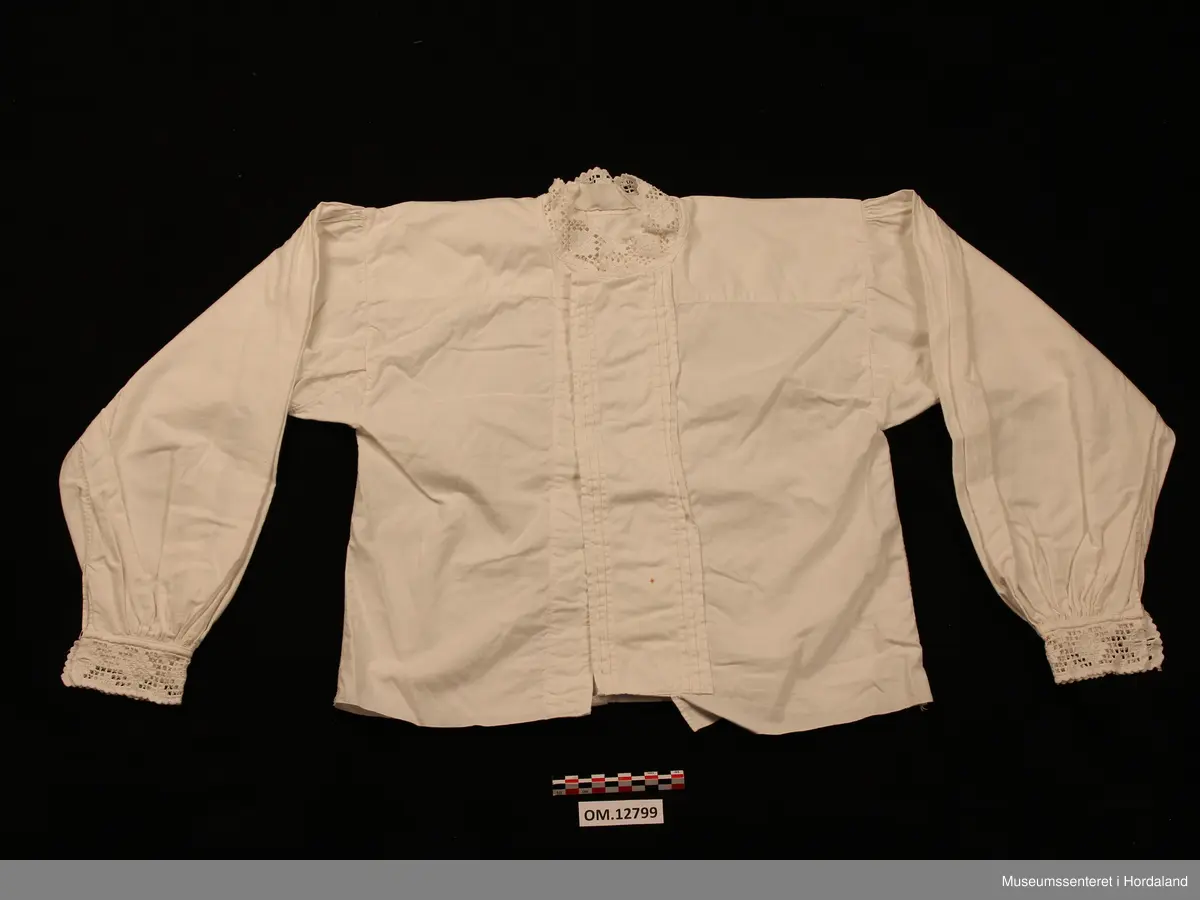 Bunadskjorte i bomullslerret med halskrage og handlinningar i hardangersaum. Brodert på svært tynt bomullstoff. Skjorta har isydde ermer med litt rynker på ermetoppen. Framme på ermene er det og rynker. Knapphol på ermelinngane til mansjettknappar. Midt framme er det ein brei stolpe med biser (legg) på kvar side. Ingen annan dekor på denne. Linningane er sydde på med hand etter at skjorta er heilt ferdig. Det er smale kanteband både framme på ermene og rundt halsen som dekker sårkantane. Så er hardangersaumlinningane nerast tråkla på. Ein rustflekk framme på stolpen.