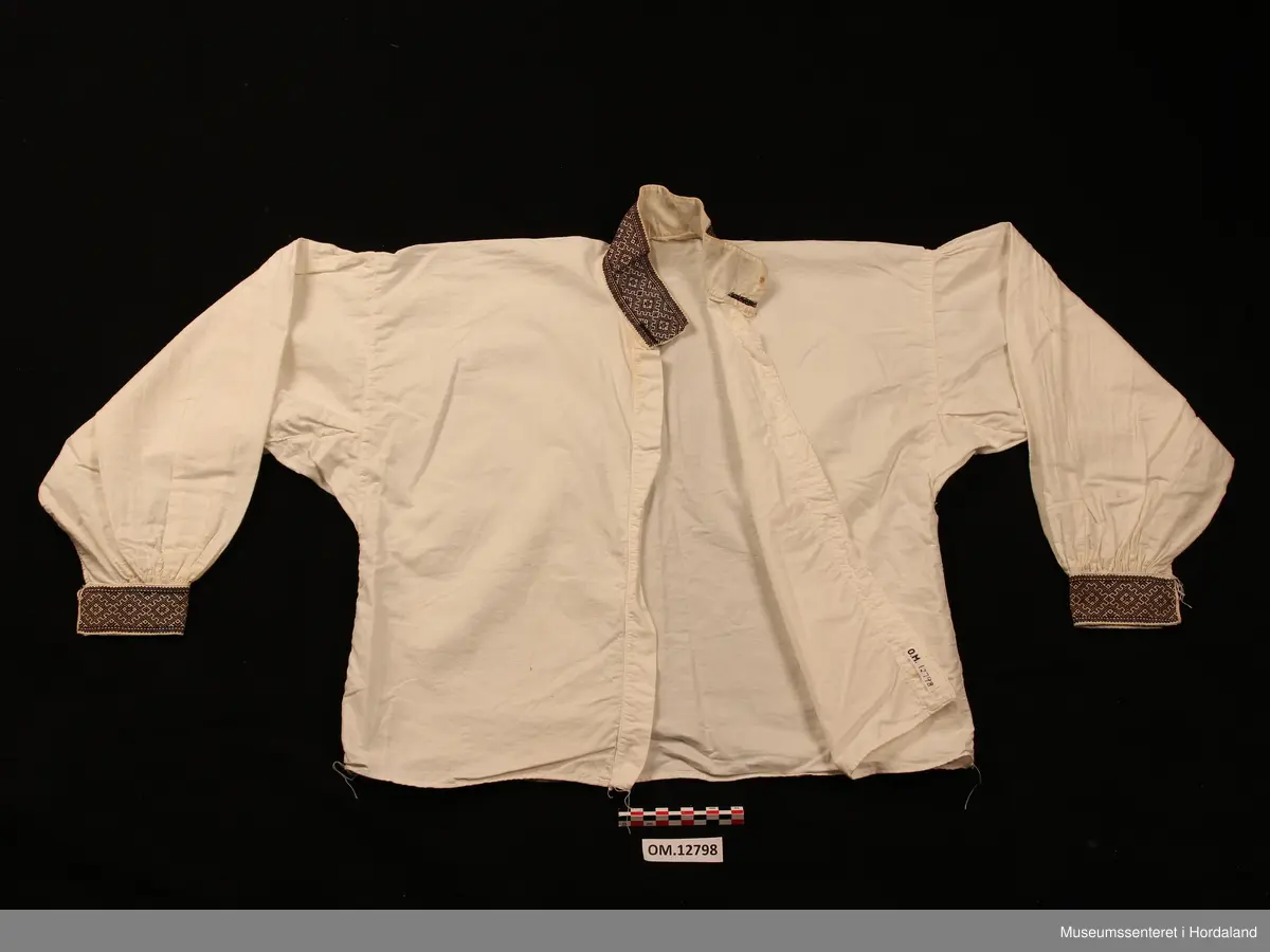 Bunadskjorte i bomullslerret med halskrage og handlinningar i smøyg (broderi). Brodert på svært tynt bomullstoff. Skjorta har isydde ermer med litt rynker på ermetoppen. Framme på ermene er det og rynker. Knapp og hempe på kvar handlinning. Stort knapphol på kvar side i halslinningen. Skjorta ber preg av at broderiet er eldre enn sjølve skjorta.
