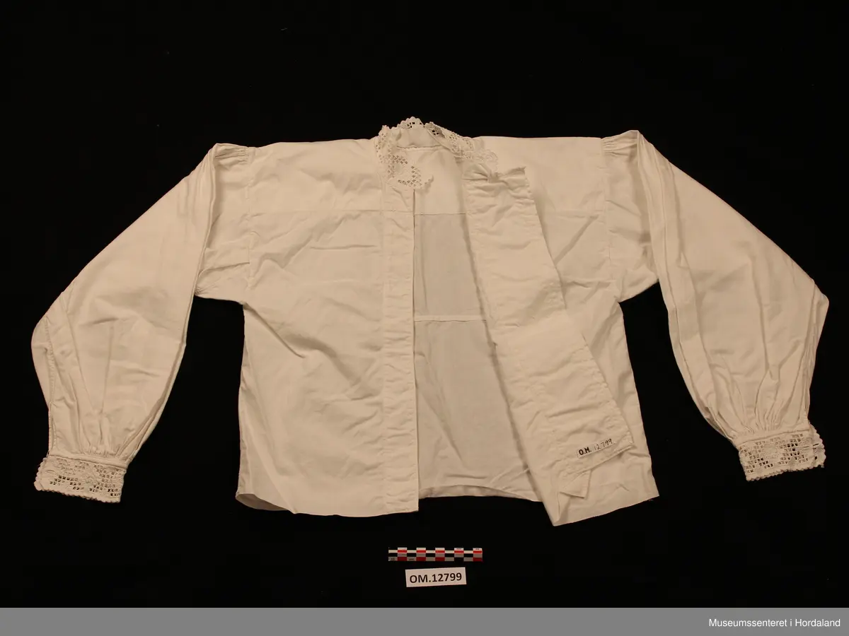 Bunadskjorte i bomullslerret med halskrage og handlinningar i hardangersaum. Brodert på svært tynt bomullstoff. Skjorta har isydde ermer med litt rynker på ermetoppen. Framme på ermene er det og rynker. Knapphol på ermelinngane til mansjettknappar. Midt framme er det ein brei stolpe med biser (legg) på kvar side. Ingen annan dekor på denne. Linningane er sydde på med hand etter at skjorta er heilt ferdig. Det er smale kanteband både framme på ermene og rundt halsen som dekker sårkantane. Så er hardangersaumlinningane nerast tråkla på. Ein rustflekk framme på stolpen.