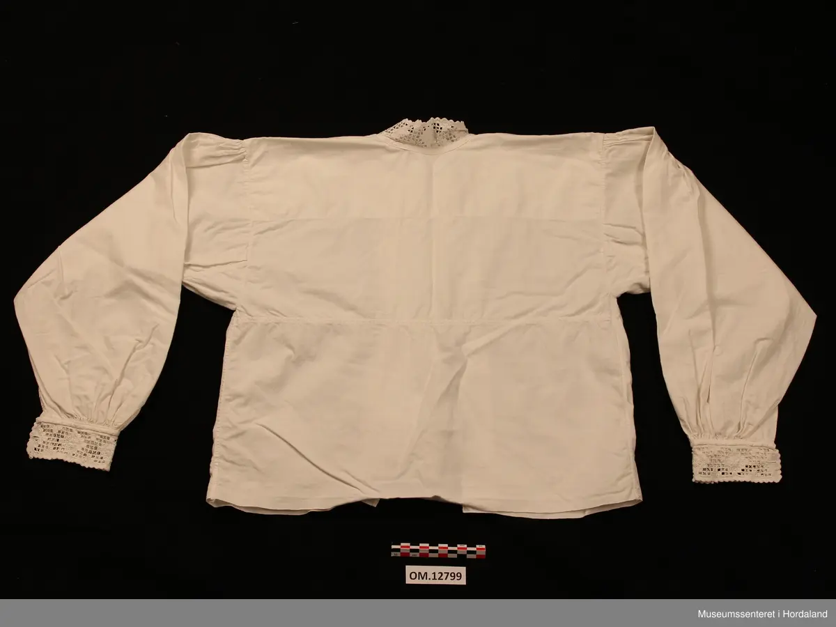 Bunadskjorte i bomullslerret med halskrage og handlinningar i hardangersaum. Brodert på svært tynt bomullstoff. Skjorta har isydde ermer med litt rynker på ermetoppen. Framme på ermene er det og rynker. Knapphol på ermelinngane til mansjettknappar. Midt framme er det ein brei stolpe med biser (legg) på kvar side. Ingen annan dekor på denne. Linningane er sydde på med hand etter at skjorta er heilt ferdig. Det er smale kanteband både framme på ermene og rundt halsen som dekker sårkantane. Så er hardangersaumlinningane nerast tråkla på. Ein rustflekk framme på stolpen.