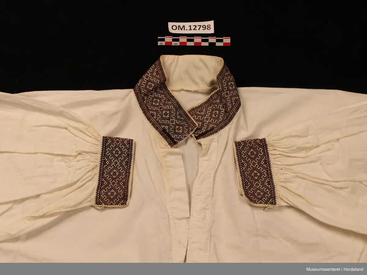 Bunadskjorte i bomullslerret med halskrage og handlinningar i smøyg (broderi). Brodert på svært tynt bomullstoff. Skjorta har isydde ermer med litt rynker på ermetoppen. Framme på ermene er det og rynker. Knapp og hempe på kvar handlinning. Stort knapphol på kvar side i halslinningen. Skjorta ber preg av at broderiet er eldre enn sjølve skjorta.